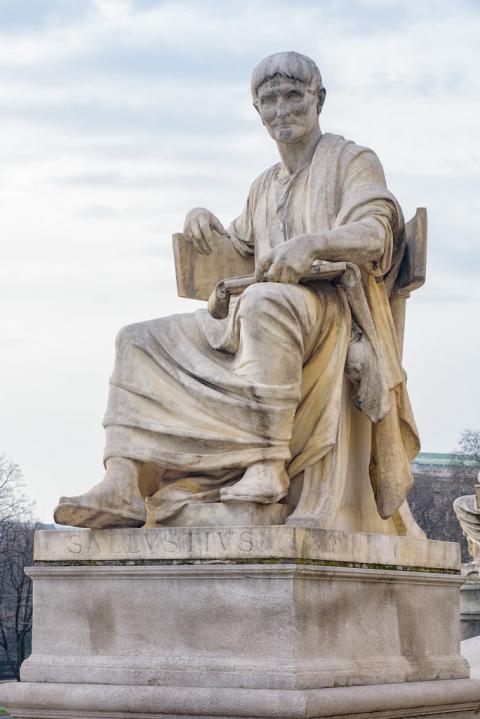 A monochromatic stone statue of a man with short hair wrapped in a toga and sitting in a large chair. His right arm is leaning on the back of the chair, and his left hand holds a writing tablet on his lap. The base of the statue reads "SALLVSTIVS"