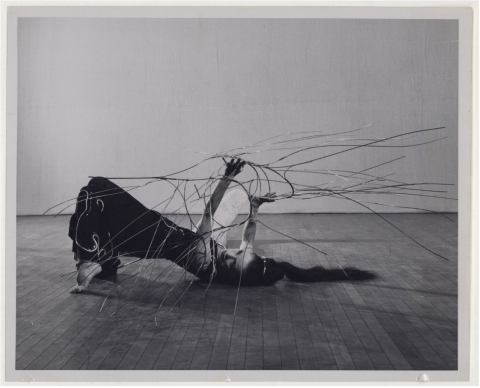 A black and white photo of a woman with long, dark hair in a ponytail lying on the floor on her tiptoes with knees bent. Her hands are raised in the air in front of her, and from her fingers many long, thin metal wires curve in different directions.