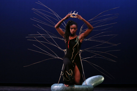 Photo of a woman standing with one foot on a smooth rock holding a crown made of bone above her head. Behind her, long sticks poke out in either direction. 