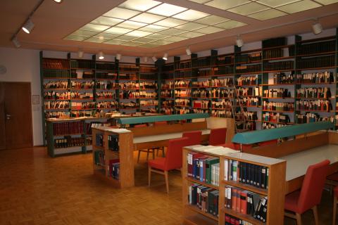 A large room with shelves of books lining the walls and wooden tables with red chairs and turquoise lamps across the middle