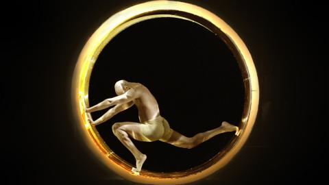 A bald, shirtless man painted gold stands at the bottom of a gold ring, pushing against one side with his legs in a lunging position