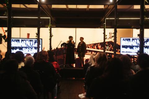 Two people stand on a stage holding microphones while an audience looks on