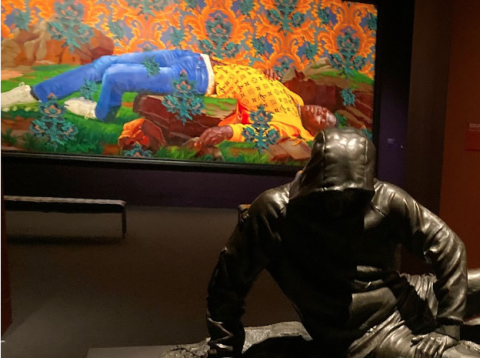 In the foreground, a bronze statue of a young person wearing jeans and hood covering their head reclines on their right arm. Behind this, a brightly-painted horizontal canvas depicts a black girl wearing a yellow printed shirt, blue jeans, and white sneakers.