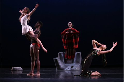 Four people stand on a dark stage. Left to right: A Black man with black straps wrapped around his torso holds a white woman in a white tutu up from her hips. They both face left, her arms extended outwards in a Y shape. Center, a white woman stands in a red and black cape with her fingertips touching at chest height. Right, a woman kneels, bending backwards, in a black dress with green and gold beads.