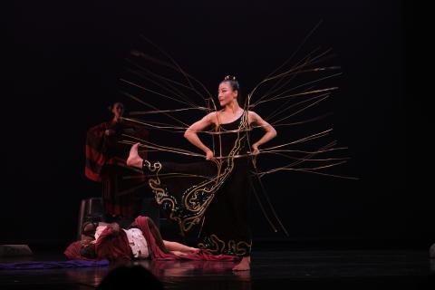 A woman in black with gold embroidery stands on the stage, her hands on her hips and one leg outstretched to the side. Long gold wires extend from all over her body.