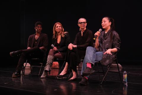 Four people sit in black folding chairs on a stage. The woman on the end speaks into a microphone.