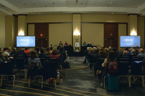 A speaker stands at a podium on a small stage while a seated audience looks on. Behind her are four panelists sitting at a table.