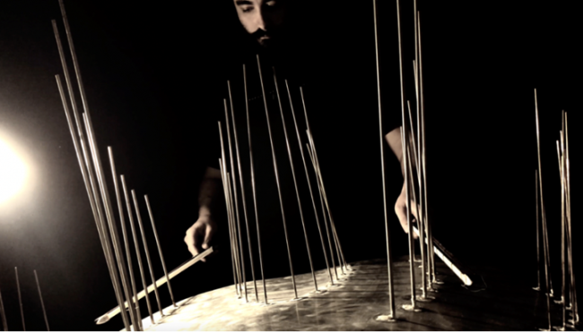 A bearded man stands in the dark over a metal plate, from which a number of vertical rods are extended. He is hitting the rods with two drumsticks.