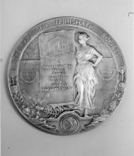 A black and white photo of a round medallion depicting a woman in a toga wearing a laurel crown holding a long papyrus scroll that reads "In commemoration of the fiftieth anniversary of the American Numismatic Society"