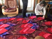 A woman in a beige dress kneels on the floor touching part of a large quilt of red and purple fabrics sewn into pointed feather shapes