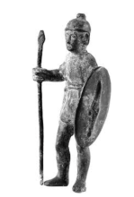 A black and white image of a stone votive figure of a man wearing a tunic and hat with a small node on the top. In his right hand, he holds a spear, and in his left hand, he holds up a round shield.
