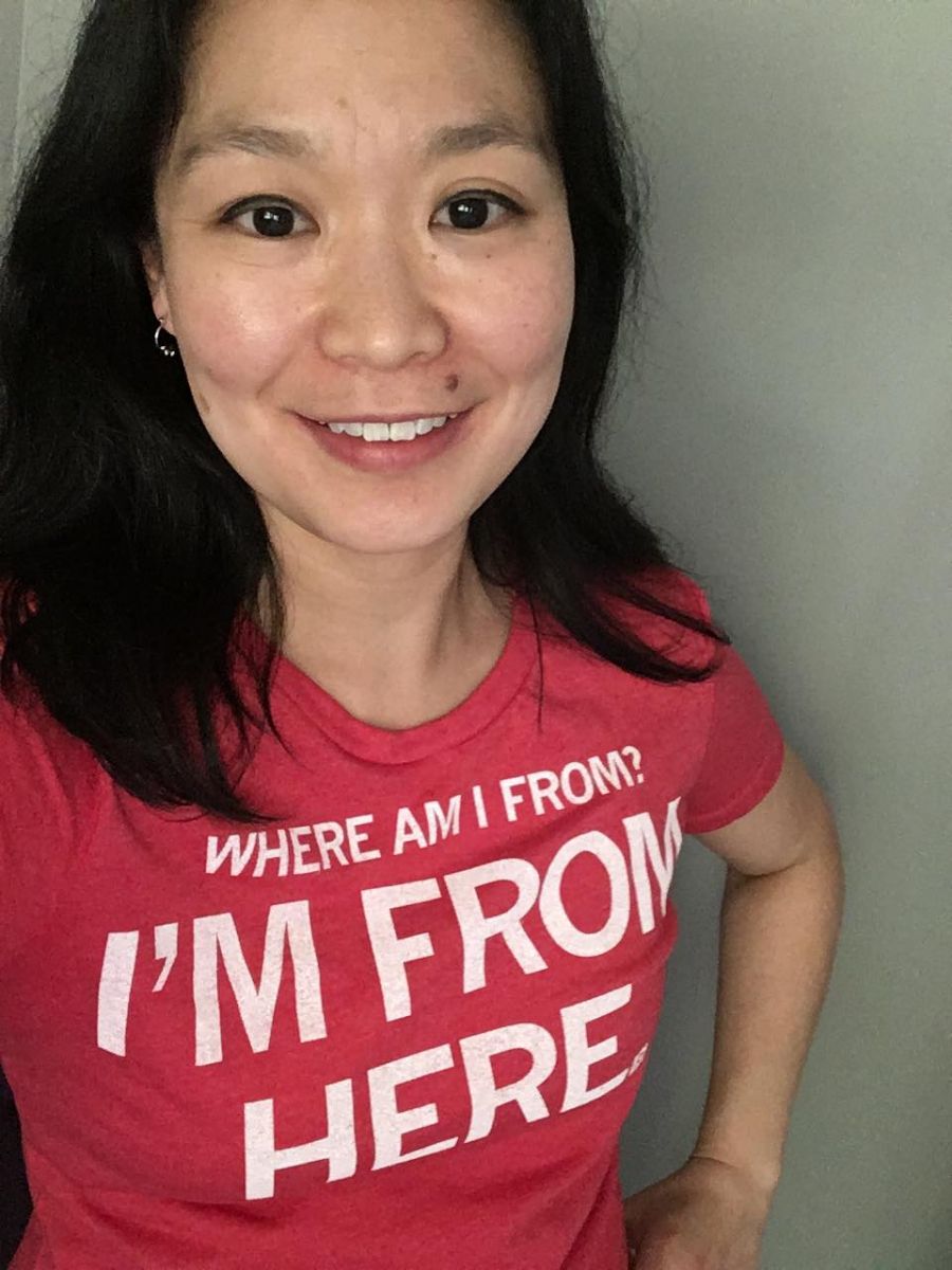 Image description: A woman smiling at the camera, wearing a red t-shirt that says "Where am I from? I'm from here."