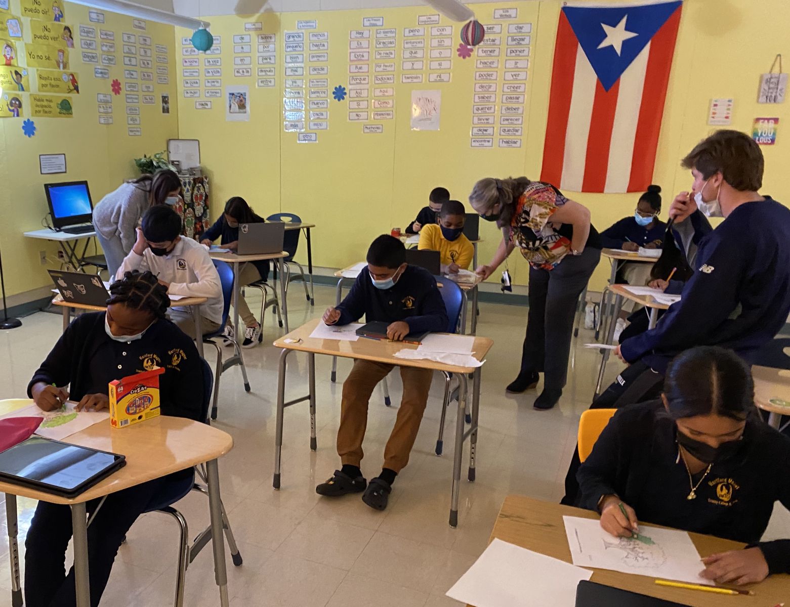 Middle schools students in uniforms sit at desks writing while teachers talk with some students individually.
