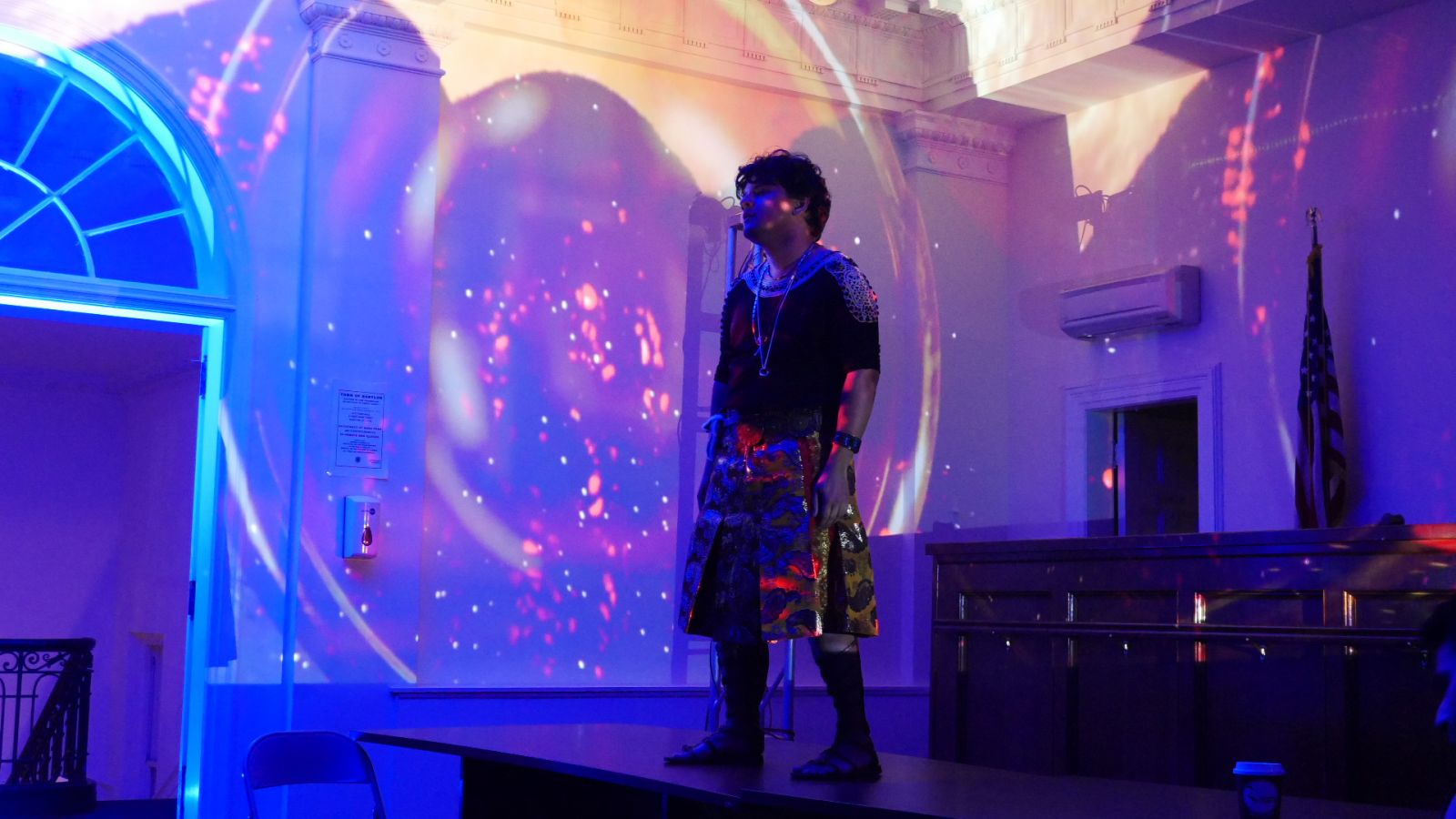 A man in a black top and gold skirt costume stands on a table in a courtroom with purple glittery lighting on the walls around him.