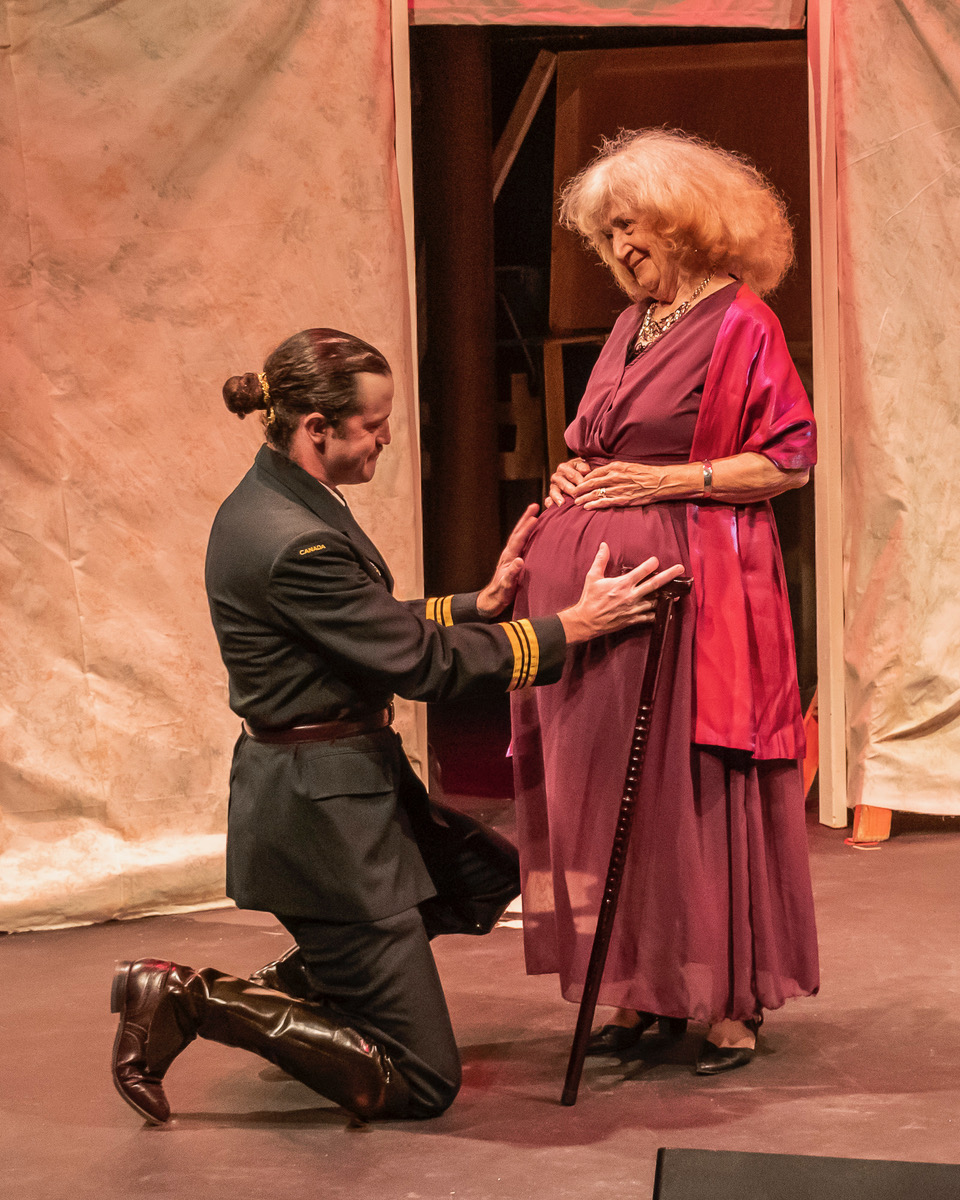 A man in military garb kneels on one knee, holding the pregnant belly of an older woman with white hair in a mauve dress.