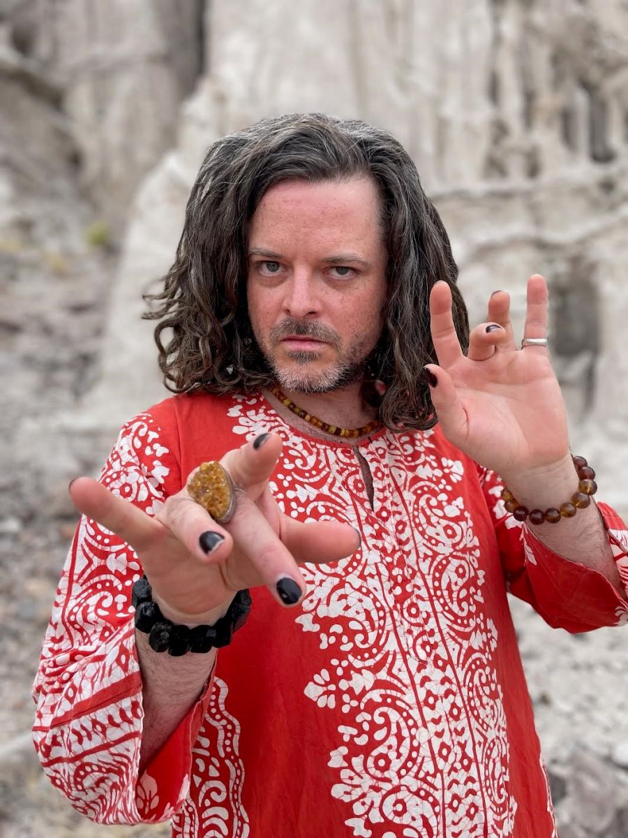 A man with shoulder-length brown hair stars at the camera. He wears a red tunic with white designs, his nails are painted, and he wiggles his fingers at the camera while they are adorned with rings and bracelets.