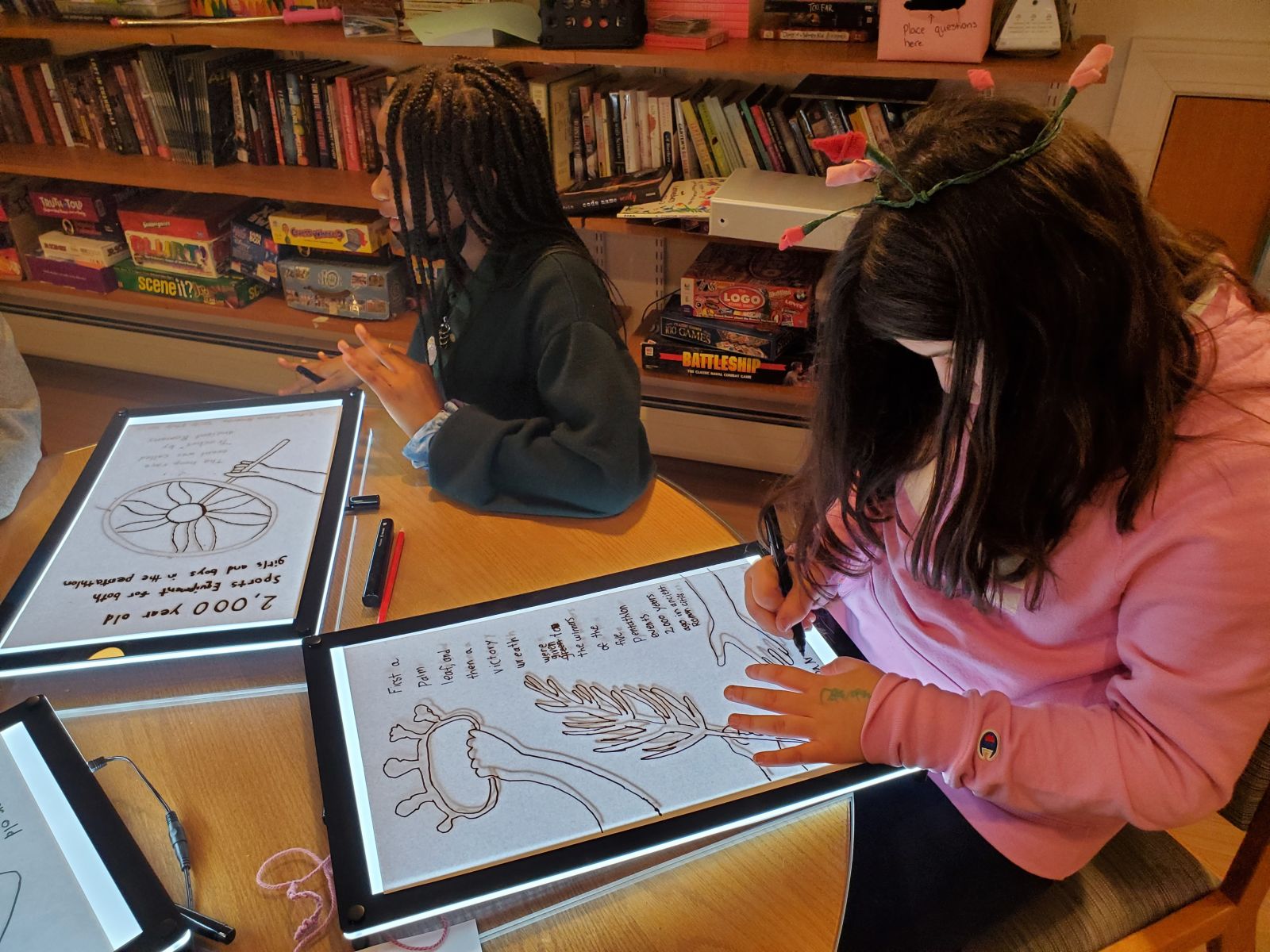 Two girls sitting at a table writing on tablets. On the left is a girl with braided hair, and on the right is a girl with short brown hair and a pink hoodie.