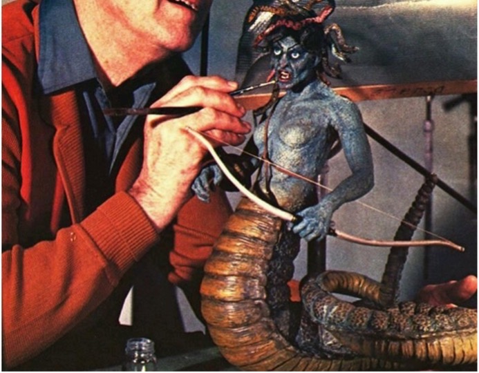A white man in a blue shirt and red cardigan painting a blue shirtless figure with horns and a brown, segmented tail