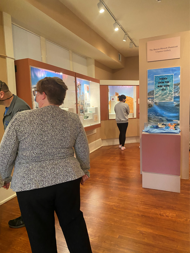 A brightly-lit indoor museum exhibit with wood floors and exhibitions on the walls. A white woman with short hair and glasses faces one of the exhibits, wearing black pants and a black and white patterned cardigan.