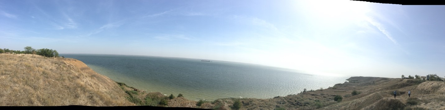 A panorama showing a dry landscape on either side of a gulf of blue ocean. There are bushes scattered around, and the sky is blue with a few clouds.